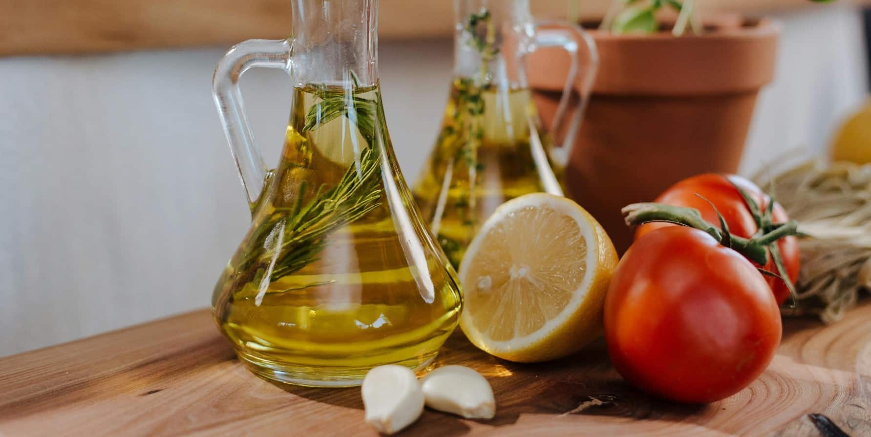 A Chopping Board with a decorative glass jug containing some herbs and Gourmet Classics White Wine Vinegar. In front is Garlic Cloves half a Lemon & 2 omatoes